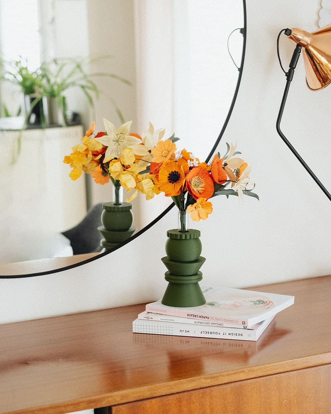 Bouquet de fleurs en papier Masterpiece Orange dans son vase vert kaki posé sur des livres sur une commode, reflet dans un grand miroir rond, décoration artisanale