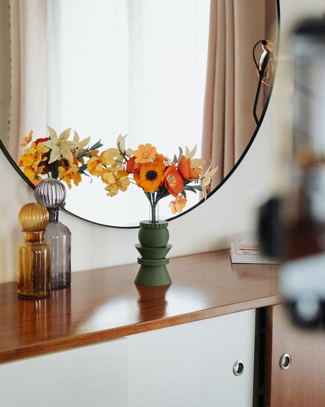 Bouquet de fleurs en papier Masterpiece Orange posé dans un vase vert kaki sur une commode vintage, avec reflet dans un miroir rond, décoration intérieure