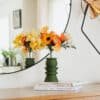 Double reflet du bouquet Masterpiece Orange dans un miroir rond, posé sur des livres sur une commode en bois, décoration intérieure artisanale