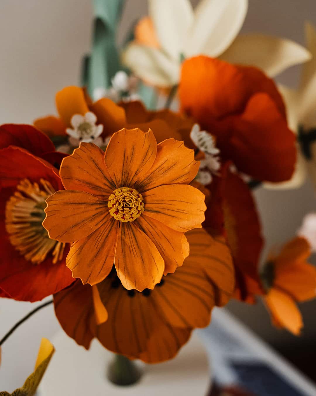 Gros plan sur un cosmos coloris clémentine en papier crépon au cœur d'un bouquet artisanal, entouré de pavots et anémones, Anaïs LVDX