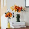 Deux bouquets de fleurs en papier de tailles différentes côte à côte sur une commode : le grand Masterpiece Orange dans un vase vert et le Garrigue dans un vase jaune