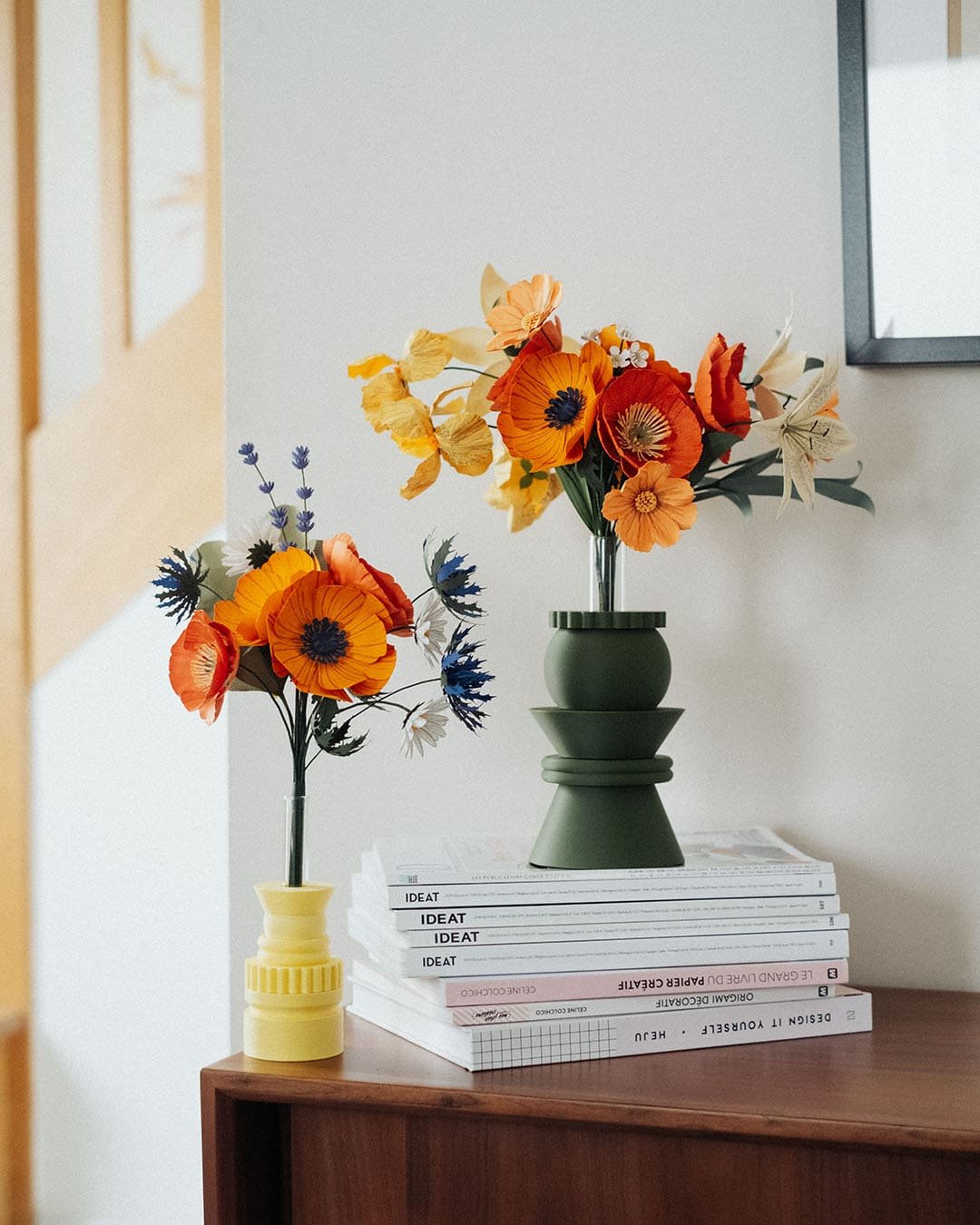 Deux bouquets de fleurs en papier de tailles différentes côte à côte sur une commode : le grand Masterpiece Orange dans un vase vert et le Garrigue dans un vase jaune