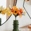 Bouquet artisanal de fleurs en papier orange jaune et ivoire dans un vase vert kaki posé sur des livres, avec lampe cuivrée et miroir rond, ambiance slow living