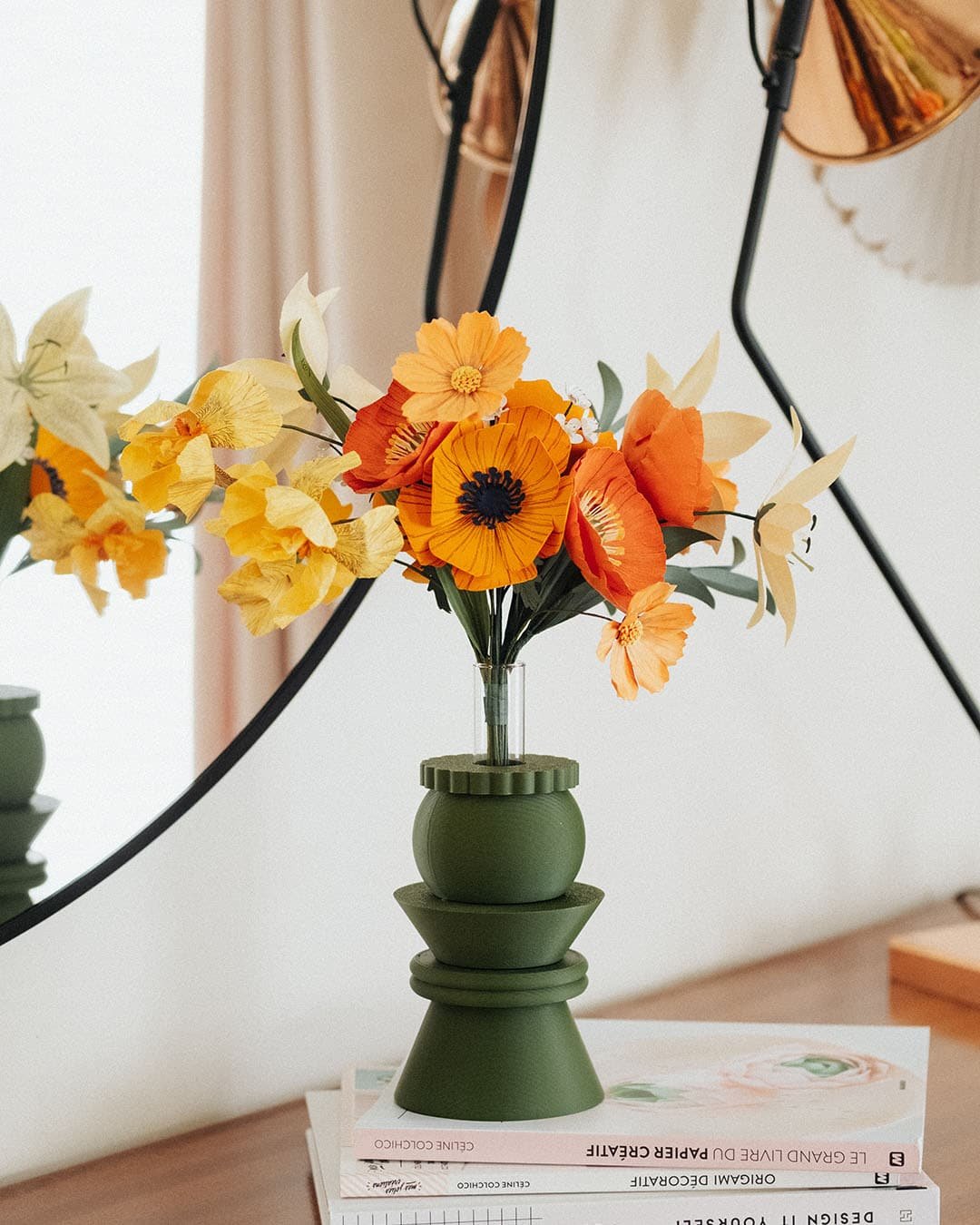Bouquet artisanal de fleurs en papier orange jaune et ivoire dans un vase vert kaki posé sur des livres, avec lampe cuivrée et miroir rond, ambiance slow living