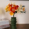 Bouquet de fleurs en papier Masterpiece Orange avec lys ivoire tacheté, cosmos clémentine et pavots orange dans un vase vert kaki, sur commode avec livres