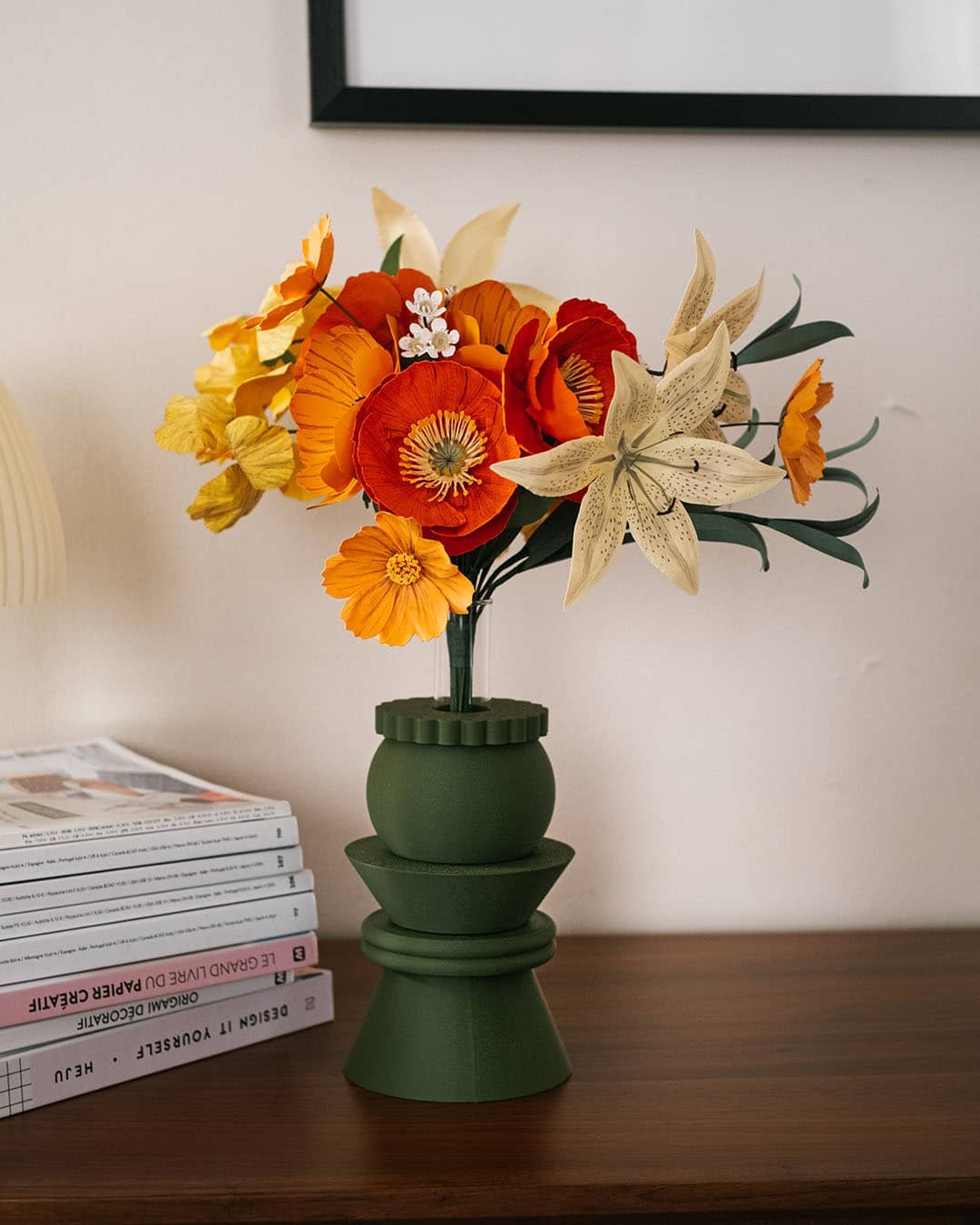 Bouquet de fleurs en papier Masterpiece Orange avec lys ivoire tacheté, cosmos clémentine et pavots orange dans un vase vert kaki, sur commode avec livres