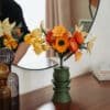 Bouquet artisanal de fleurs en papier orange et jaune dans un vase vert kaki géométrique posé sur une commode en bois, reflet miroir en arrière-plan