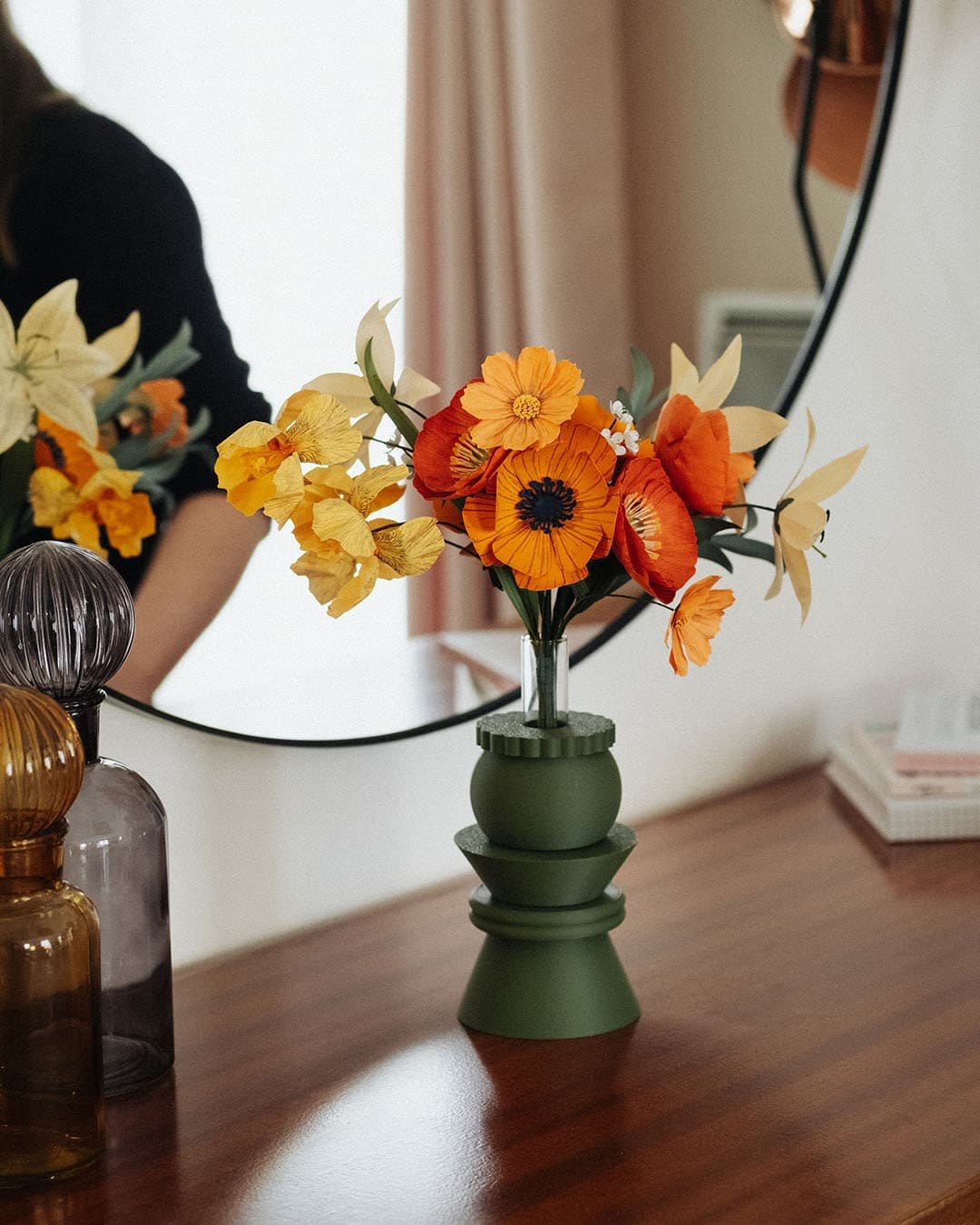 Bouquet artisanal de fleurs en papier orange et jaune dans un vase vert kaki géométrique posé sur une commode en bois, reflet miroir en arrière-plan