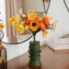 Bouquet de fleurs en papier orange jaune et ivoire dans un vase vert géométrique posé sur des livres, devant un miroir rond, décoration intérieure slow living