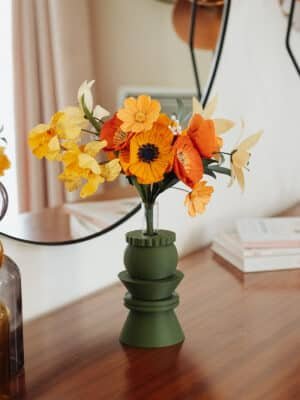 Bouquet de fleurs en papier orange jaune et ivoire dans un vase vert géométrique posé sur des livres, devant un miroir rond, décoration intérieure slow living