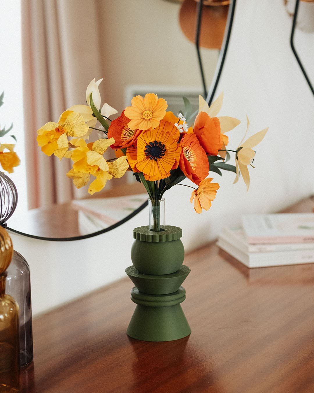 Bouquet de fleurs en papier orange jaune et ivoire dans un vase vert géométrique posé sur des livres, devant un miroir rond, décoration intérieure slow living