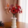 Bouquet artisanal de fleurs en papier d'art rouge orange et rose dans un vase blanc posé sur une commode en bois, rideau rose poudré en arrière-plan