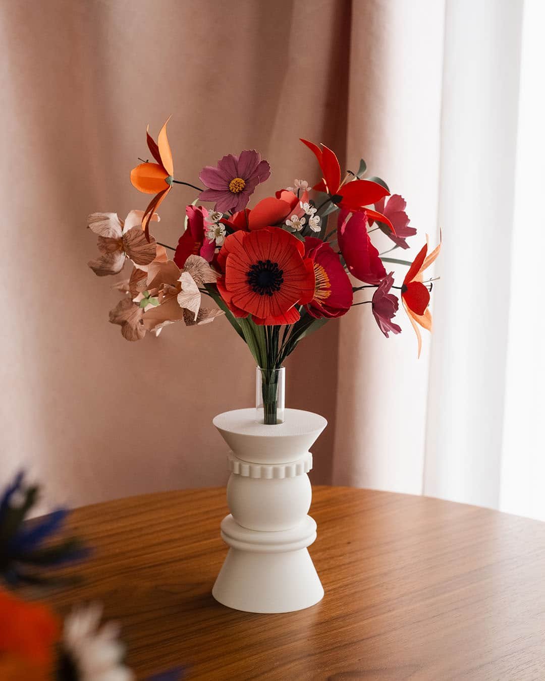Bouquet artisanal de fleurs en papier d'art rouge orange et rose dans un vase blanc posé sur une commode en bois, rideau rose poudré en arrière-plan