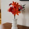 Bouquet de fleurs en papier d'art avec lys orange tacheté dominant la composition, cosmos hortensia et pavots fuchsia, dans un vase blanc sur commode en bois