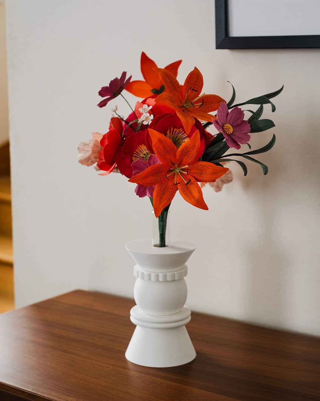 Bouquet de fleurs en papier d'art avec lys orange tacheté dominant la composition, cosmos hortensia et pavots fuchsia, dans un vase blanc sur commode en bois