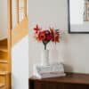 Bouquet de fleurs en papier d'art Masterpiece Rose dans un vase blanc sur commode, avec escalier en bois et poster botanique encadré en arrière-plan