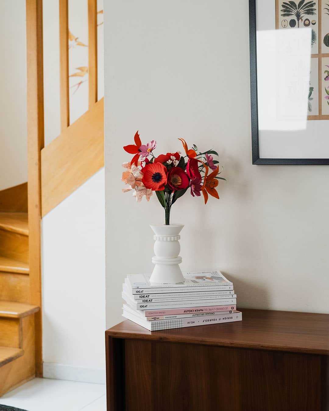 Bouquet de fleurs en papier d'art Masterpiece Rose dans un vase blanc sur commode, avec escalier en bois et poster botanique encadré en arrière-plan