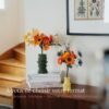 Deux bouquets artisanaux de fleurs en papier posés sur une commode : grand bouquet Masterpiece Orange dans un vase vert kaki et petit bouquet Garrigue dans un vase jaune, escalier en bois visible