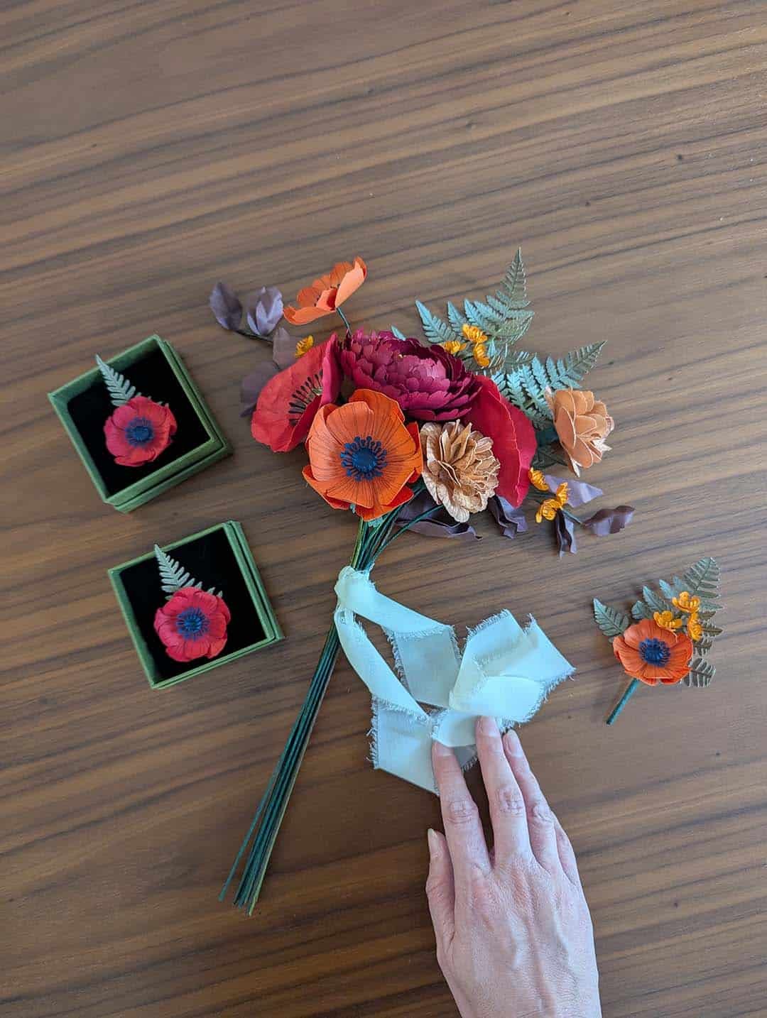 Bouquet de mariée fleurs en papier rouge, orange et lie de vin en cours de finition avec ruban et broches assorties