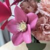 Gros plan sur une hellébore viola et des œillets rosa en papier d'art, avec une tulipe fuchsia, dans un vase vert olive