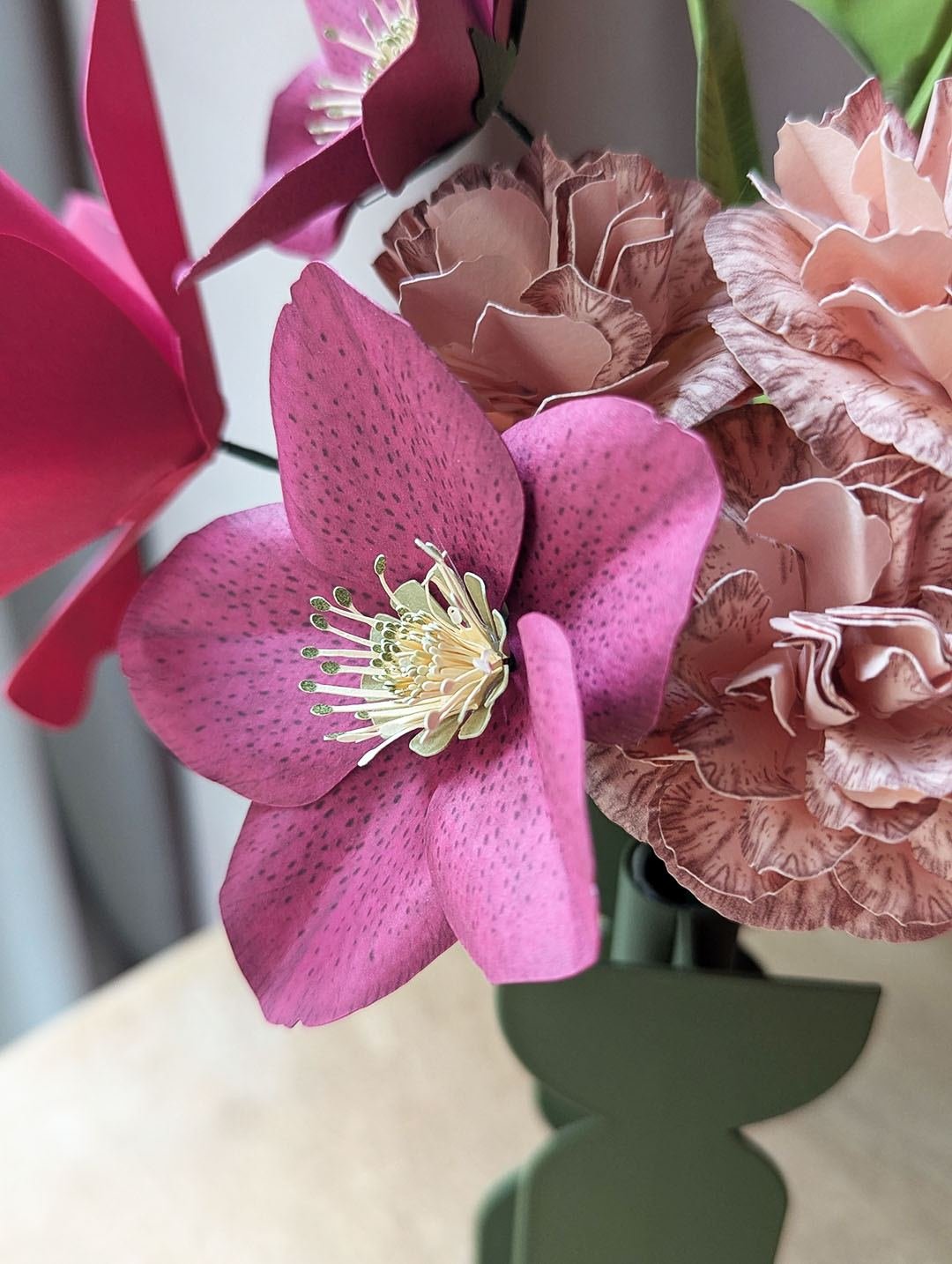 Gros plan sur une hellébore viola et des œillets rosa en papier d'art, avec une tulipe fuchsia, dans un vase vert olive