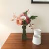 Bouquet de fleurs en papier rose, blanc et vert dans un vase olive posé près de deux pots en céramique blanche