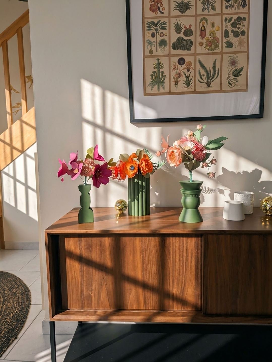 Trois bouquets de fleurs en papier fuchsia, orange et rosa dans des vases vert olive sur un buffet en noyer, sous une planche botanique