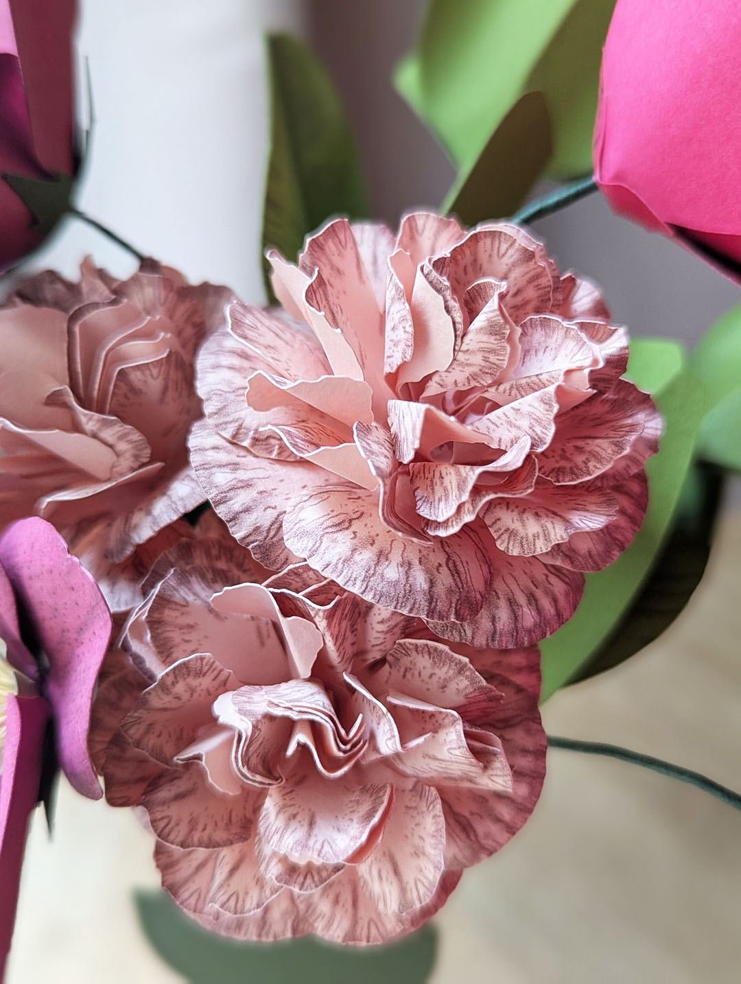 Gros plan sur des œillets rosa en papier d'art aux pétales froissés et veinés, entourés de feuillages pistache