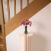 Bouquet de fleurs en papier fuchsia et rosa dans un vase vert olive sur un socle plissé, devant un escalier en bois
