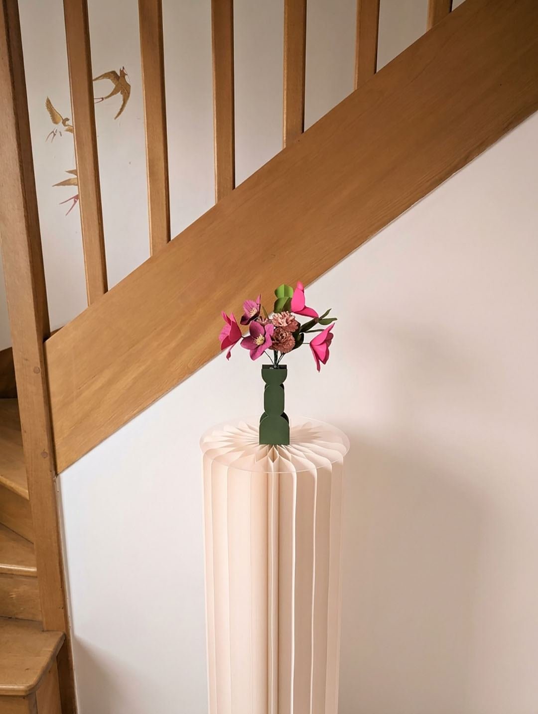 Bouquet de fleurs en papier fuchsia et rosa dans un vase vert olive sur un socle plissé, devant un escalier en bois