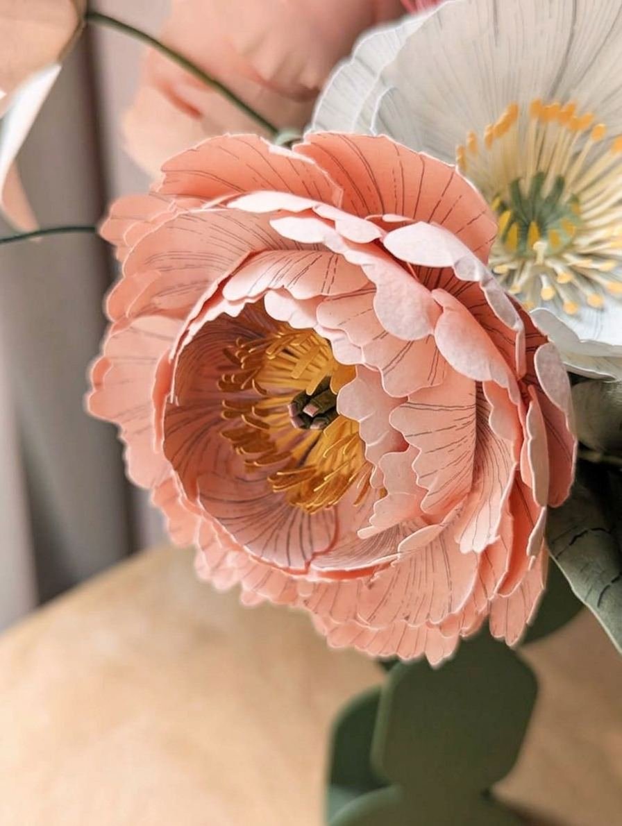 Gros plan sur une pivoine rosa en papier d'art avec étamines jaunes, à côté d'un pavot blanc