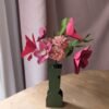 Bouquet de fleurs en papier fuchsia, viola et rosa dans un vase vert olive sur une table en bois, devant un rideau rose
