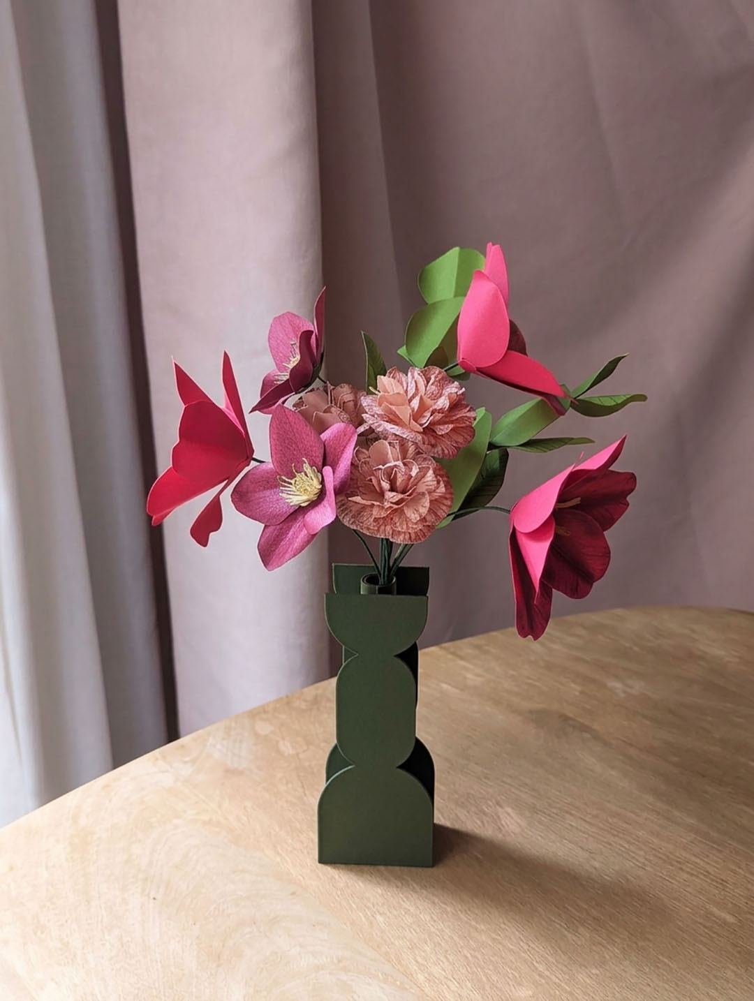 Bouquet de fleurs en papier fuchsia, viola et rosa dans un vase vert olive sur une table en bois, devant un rideau rose