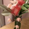 Bouquet de fleurs en papier avec un leucospermum rose vénitien, des trèfles rosa et un vase vert olive, devant un rideau rose