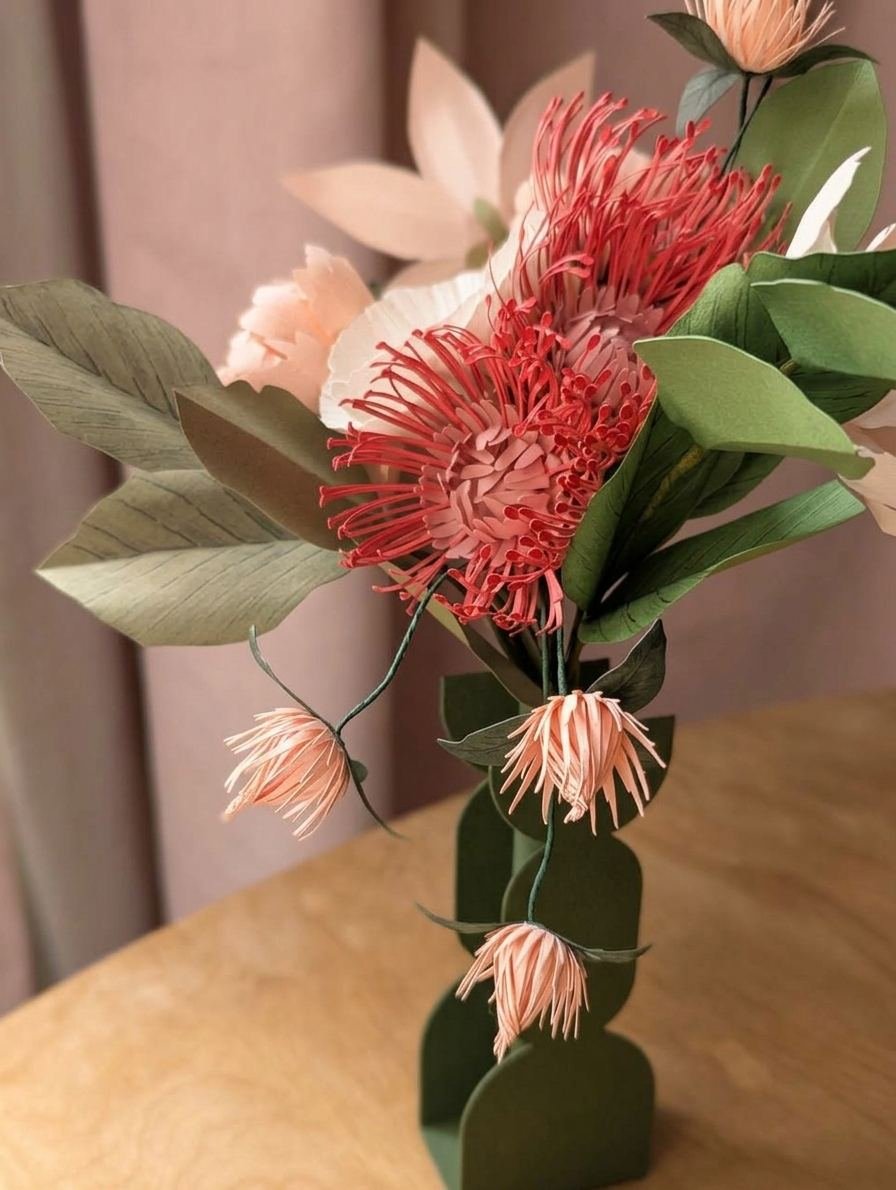 Bouquet de fleurs en papier avec un leucospermum rose vénitien, des trèfles rosa et un vase vert olive, devant un rideau rose