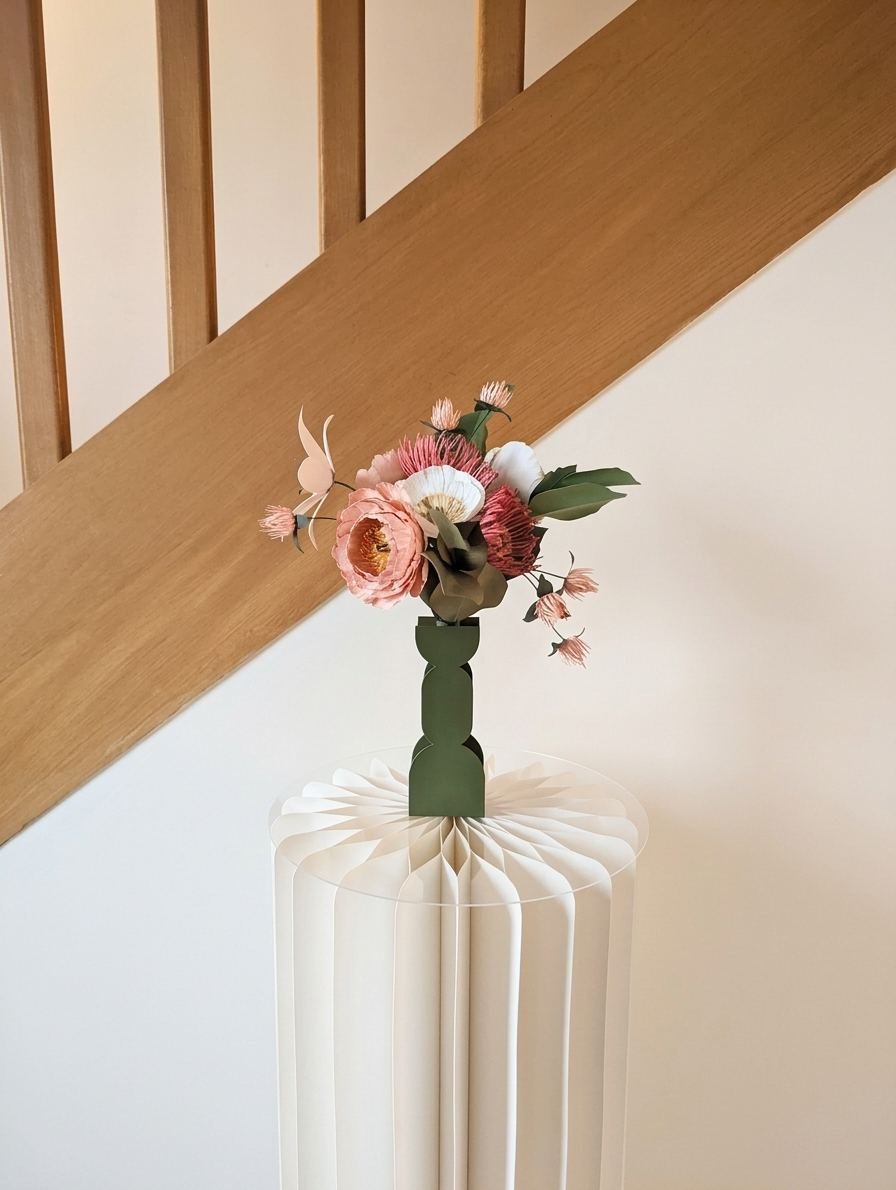Bouquet de fleurs en papier dans un vase vert olive posé sur un socle en papier plissé devant un escalier en bois