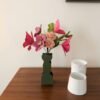 Bouquet de fleurs en papier fuchsia dans un vase vert olive sur une table en noyer, près de deux pots en céramique blanche