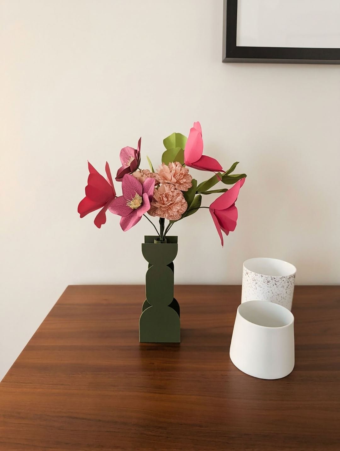 Bouquet de fleurs en papier fuchsia dans un vase vert olive sur une table en noyer, près de deux pots en céramique blanche