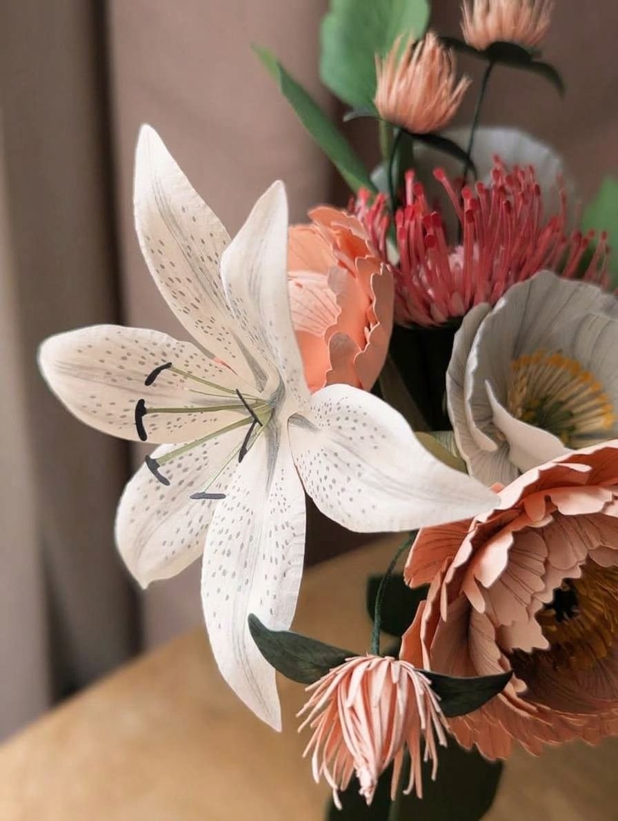 Gros plan sur un lys aurore blanc moucheté en papier d'art, entouré de pivoines et d'un pavot en papier