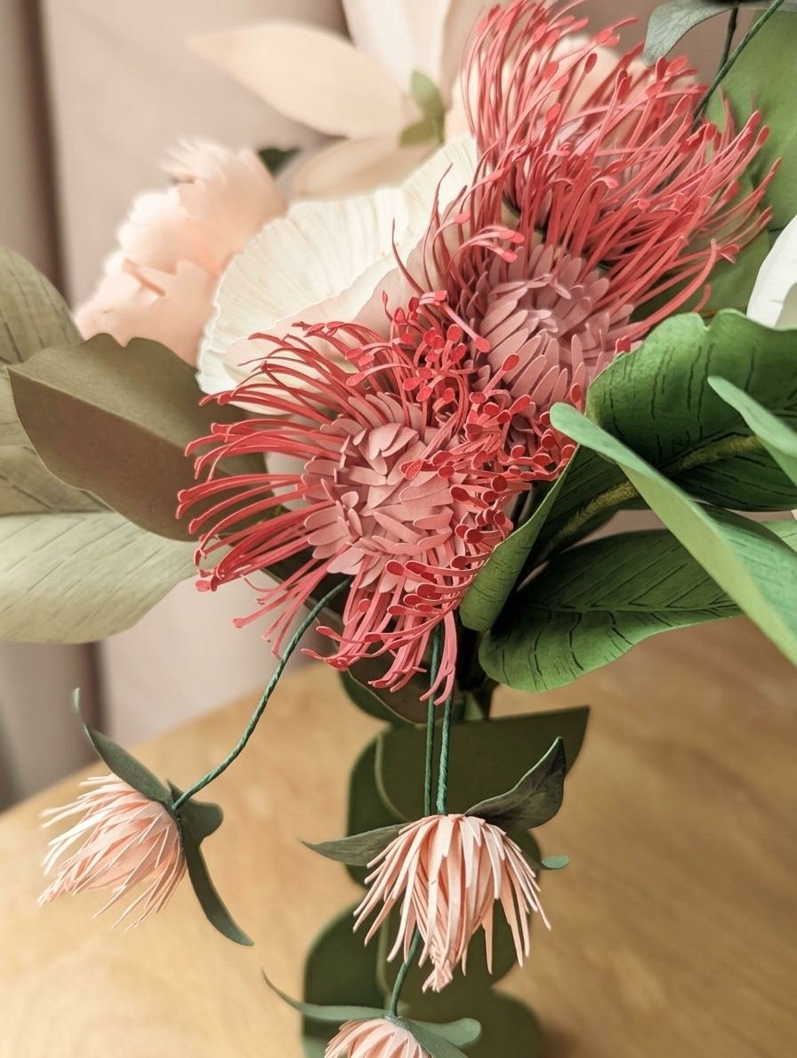 Gros plan sur des leucospermums rose vénitien et des fleurs de trèfle rosa en papier, avec feuillages de hêtre