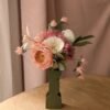 Bouquet de fleurs en papier rose, blanc et vert olive dans un vase graphique, devant un rideau rose poudré