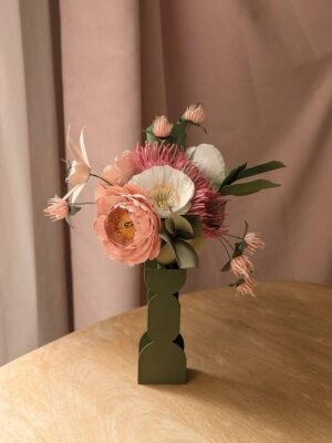 Bouquet de fleurs en papier rose, blanc et vert olive dans un vase graphique, devant un rideau rose poudré