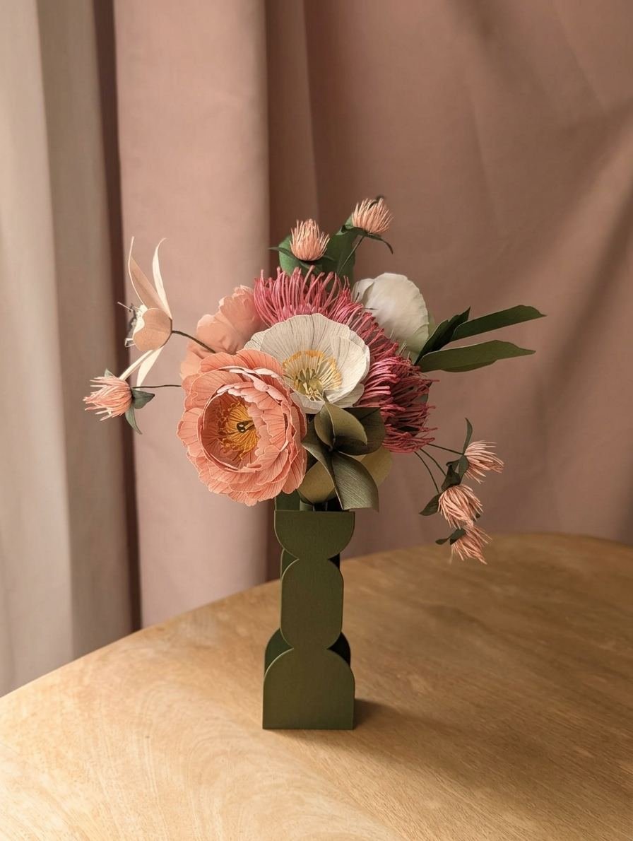 Bouquet de fleurs en papier rose, blanc et vert olive dans un vase graphique, devant un rideau rose poudré