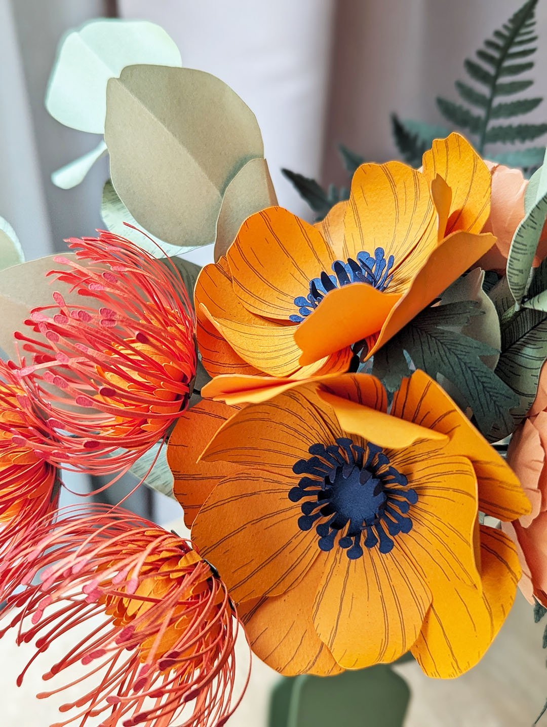 Détail de deux anémones jaune soleil en papier au cœur bleu nuit, entourées de leucospermums et de grandes feuilles d'eucalyptus
