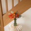 Bouquet de fleurs en papier orange dans un vase vert LA CHAMBRE D'AMI posé sur une colonne en papier plissé devant un escalier en bois