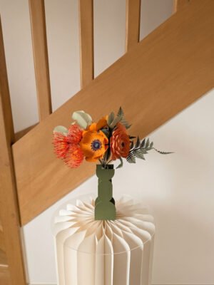 Bouquet de fleurs en papier orange dans un vase vert LA CHAMBRE D'AMI posé sur une colonne en papier plissé devant un escalier en bois