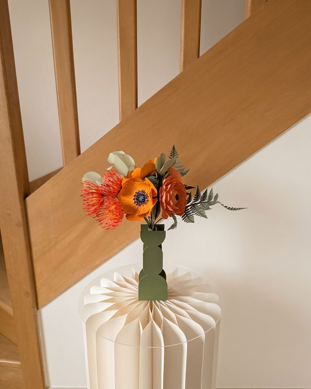 Bouquet de fleurs en papier orange dans un vase vert LA CHAMBRE D'AMI posé sur une colonne en papier plissé devant un escalier en bois