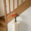 Bouquet de fleurs en papier Théodore dans un vase vert sur une colonne blanche plissée, à côté d'un escalier en chêne