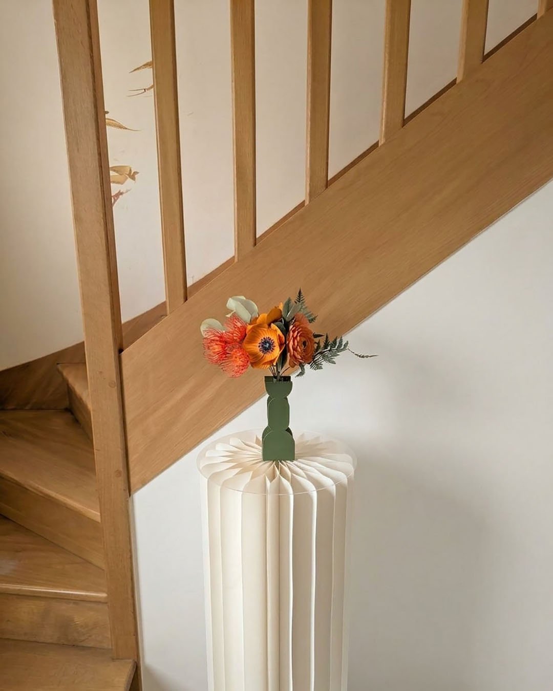 Bouquet de fleurs en papier Théodore dans un vase vert sur une colonne blanche plissée, à côté d'un escalier en chêne
