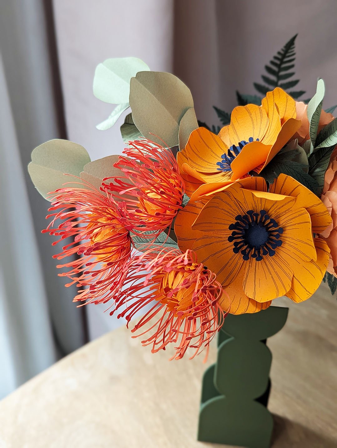 Gros plan sur trois leucospermums orange et deux anémones jaune soleil en papier d'art avec eucalyptus vert tilleul